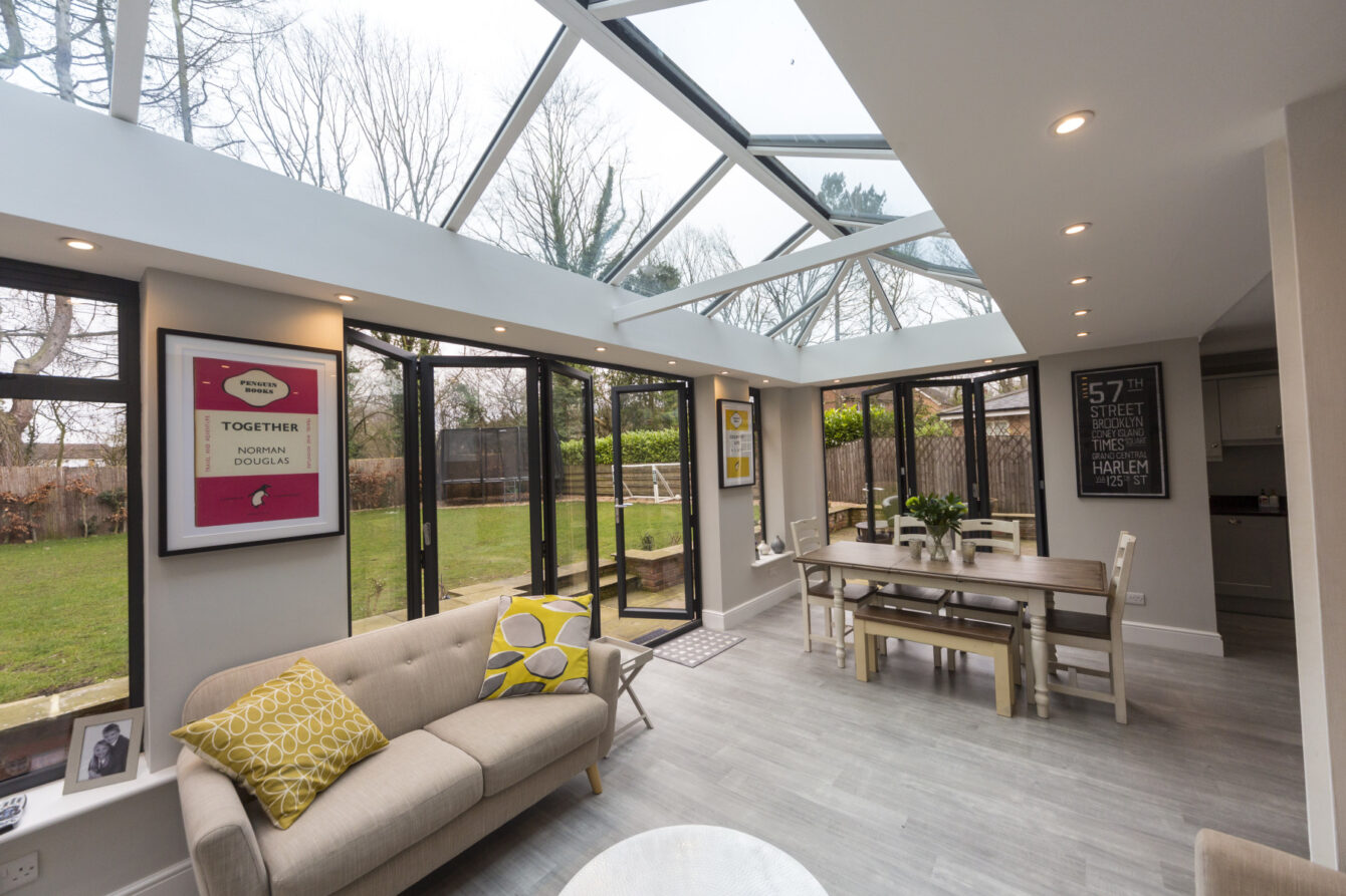A large conservatory with a dining space at one end and an area for relaxing with a sofa at the other. The roof is one long roof lantern, spotlights are set into the ceiling on either side.