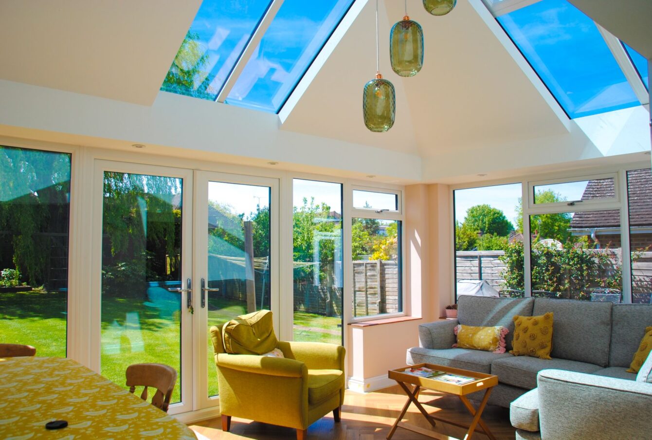 A bright corner of a conservatory with comfortable chairs and sofa. The corners of the building are solid with large central skylights, outside is a garden in summer.