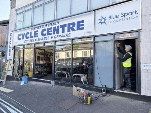 Haywards Heath Cycle Centre shopfront showing Blue Spark Organisation next door with a workman in a high-vis vest working in the doorway. 