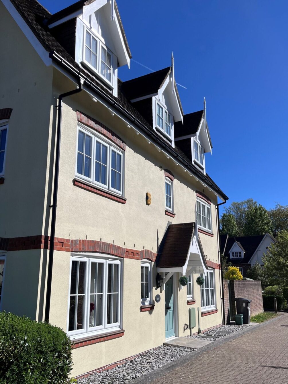 View of a three storey, double fronted house from the left. There are three pitched Dorma windows at the top with a central front door with a pitched-roof, open porch. The finish is cream render with natural brick detailing.