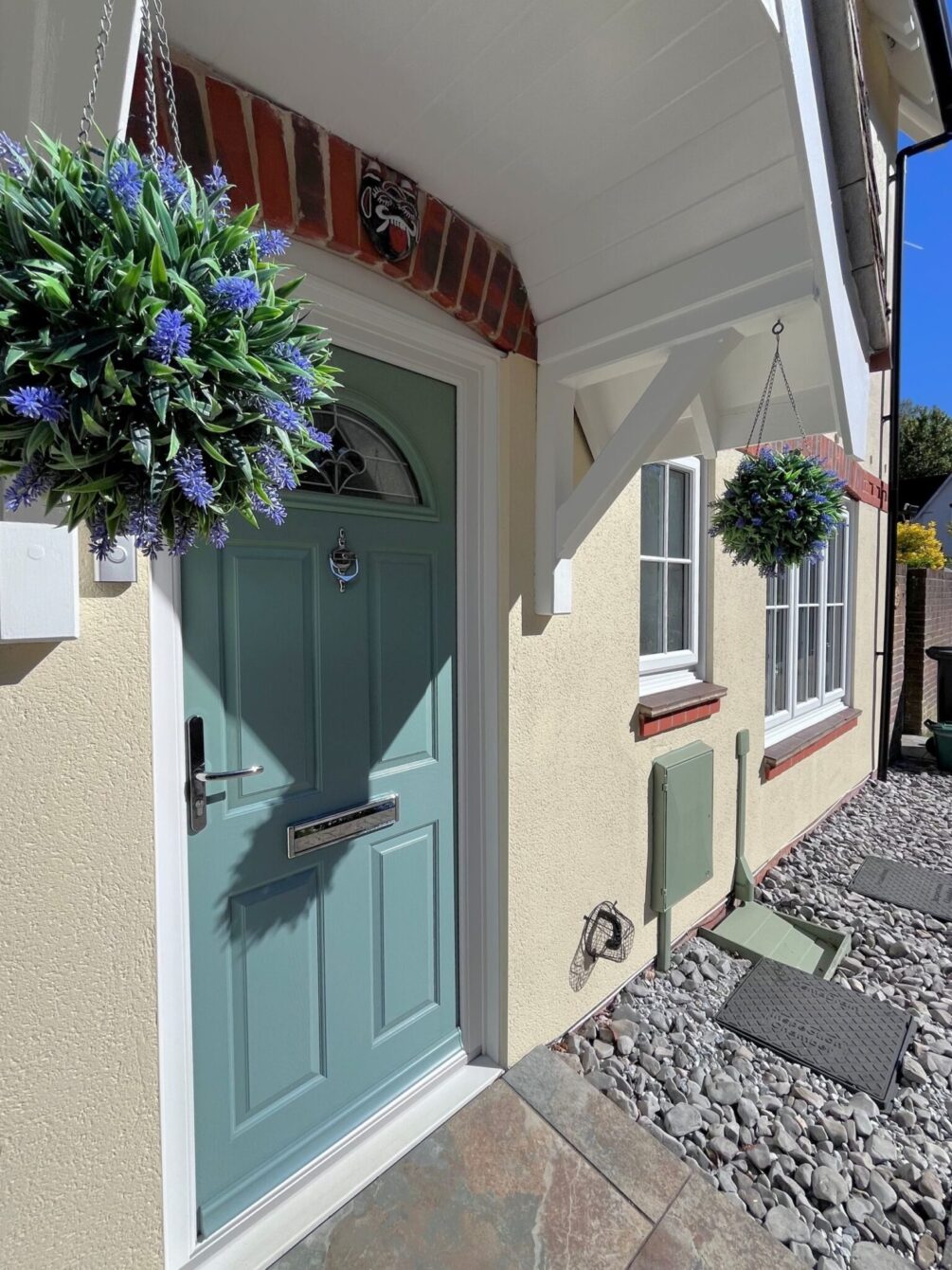 Close up of a cream rendered house with a smart, sage green Duo Composite front door. There are hanging baskets each side.