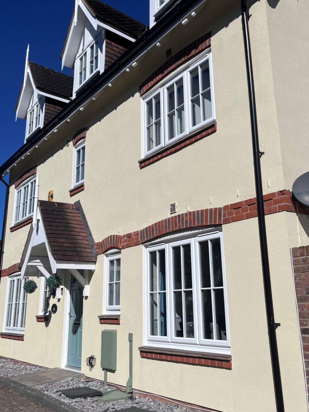 View of a three storey, double fronted house from the right. There are three pitched Dorma windows at the top with a central front door with a pitched-roof, open porch. The finish is cream render with natural brick detailing.