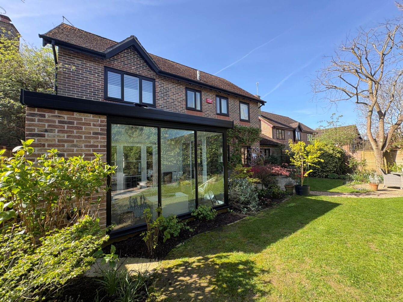 A house with a modern hup! extension to the back with three large windows and a closed-in section with brick-effect panelling to match the house. It overlooks a spacious mature garden.