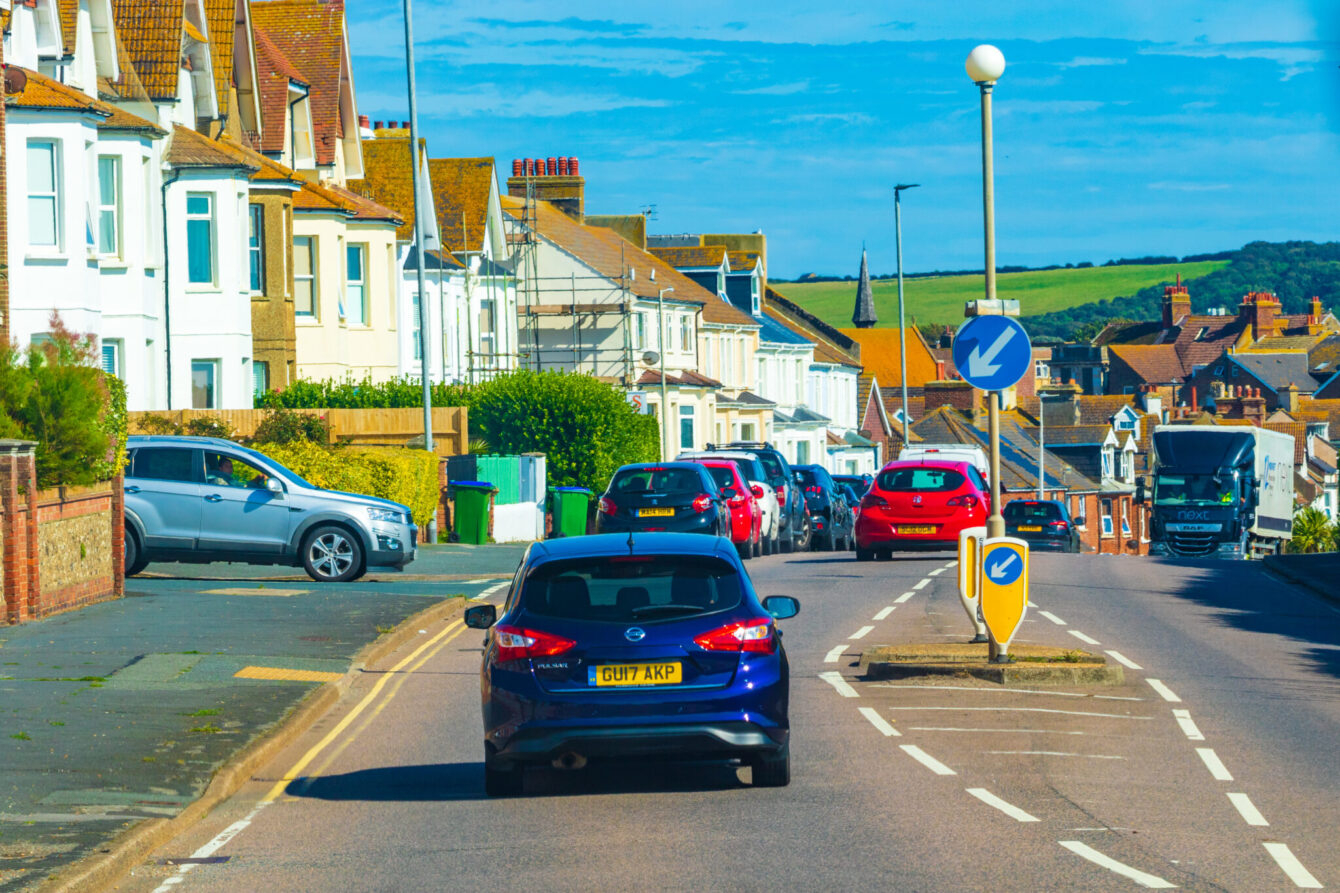 A busy road next to a row of houses with fields in the background.